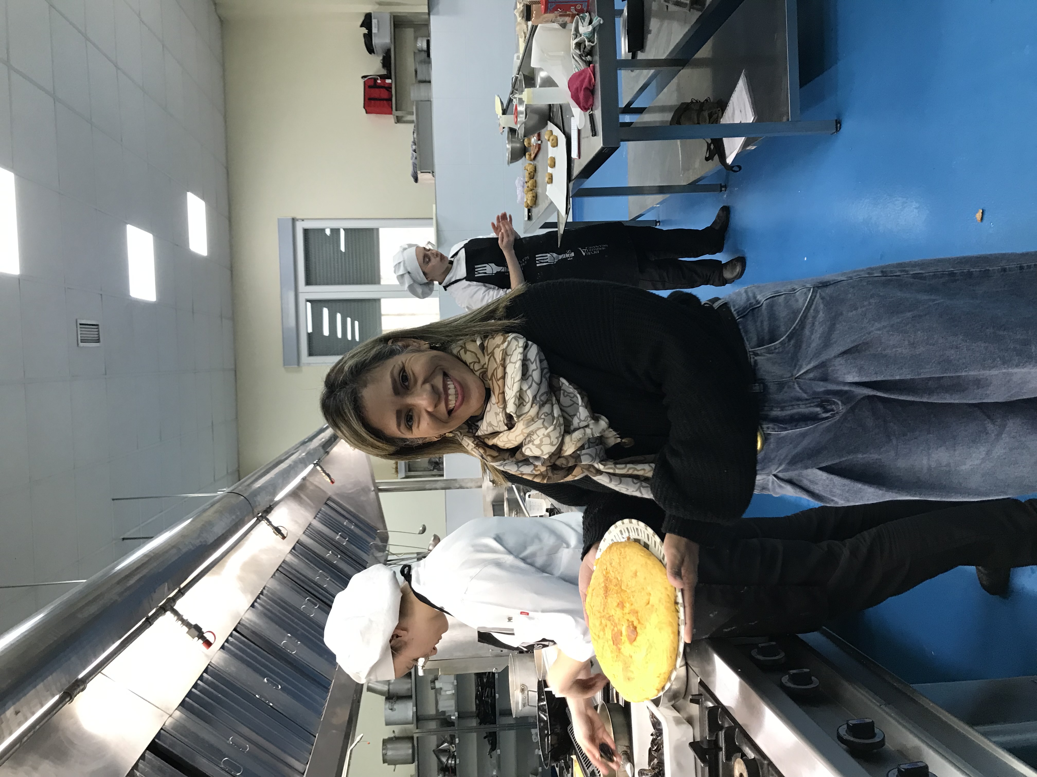 Yfigenia Moreno Aponte sonriendo en una cocina profesional, sosteniendo un plato con una tortilla recién horneada, con dos cocineras trabajando al fondo
